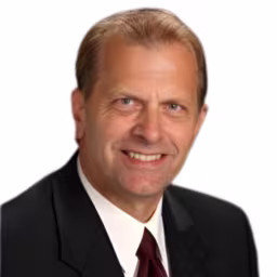 headshot of Jim Ross, wearing a black suit with a red tie