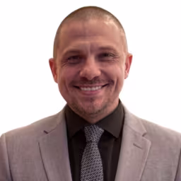 Headshot of Jimmy Litman wearing black dress shirt, black and grey tie, and grey suit coat with white background