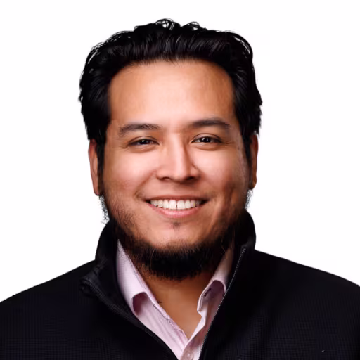 Headshot of Nick Sosa wearing pink dress shirt and black zip up sweater with white background