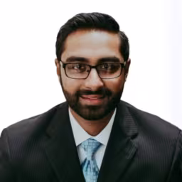 Headshot of Shiva Vasudevan wearing white dress shirt, light blue tie, and blacck suit with white background
