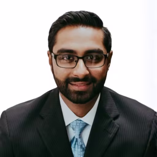 Headshot of Shiva Vasudevan wearing white dress shirt, light blue tie, and blacck suit with white background