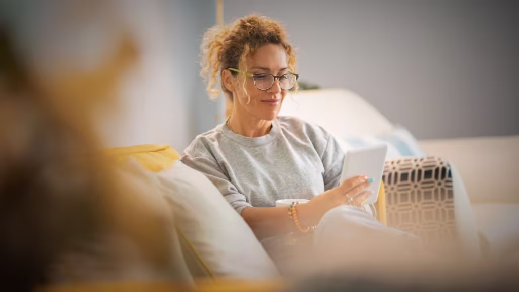 Young mature woman smile and enjoy time reading an electronic book on tablet reader device comfortably sitting on the sofa