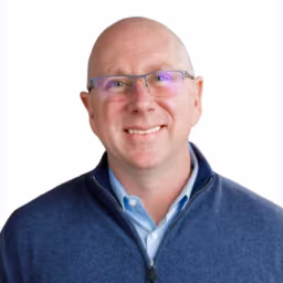 Chris Fleming wearing navy blue sweater with light blue button up shirt underneath with white background